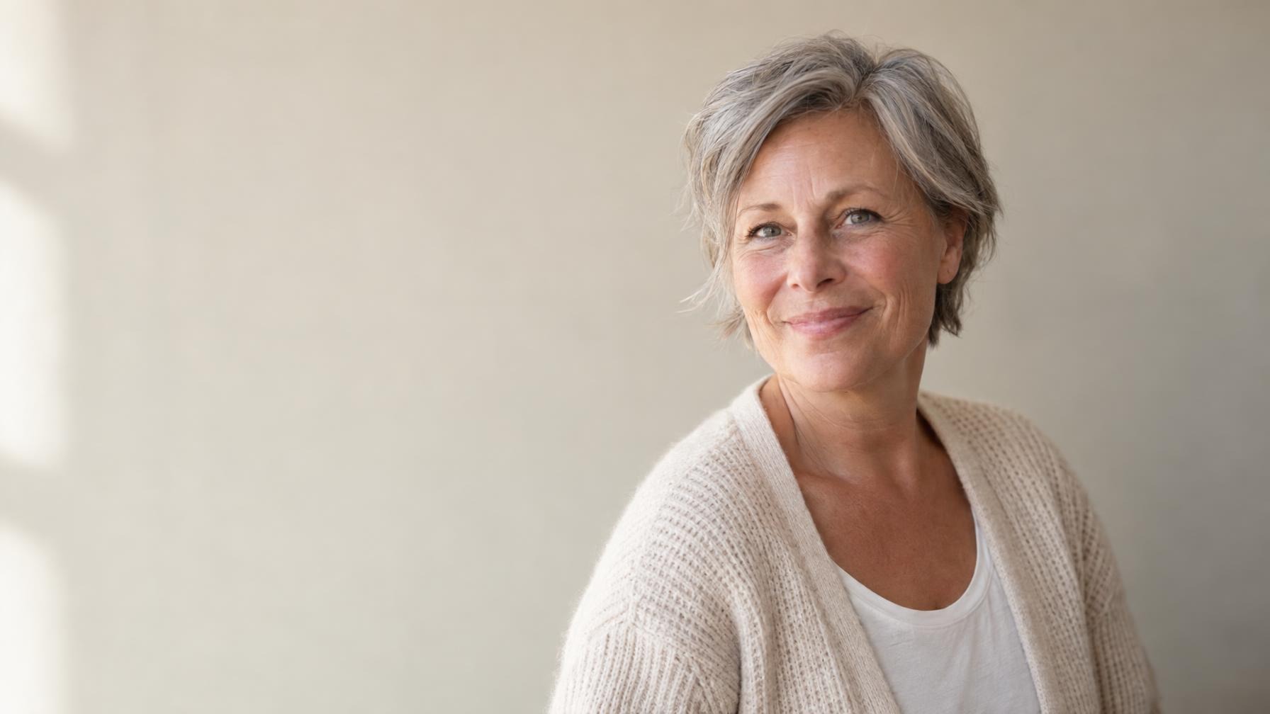 Vrouw van rond de vijftig met kort grijs haar in een crème vest tegen een rustige beige achtergrond