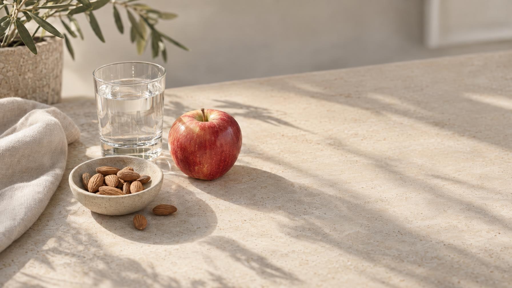 Appel, glas water en een schaaltje amandelen op een lichte stenen tafel in warm daglicht