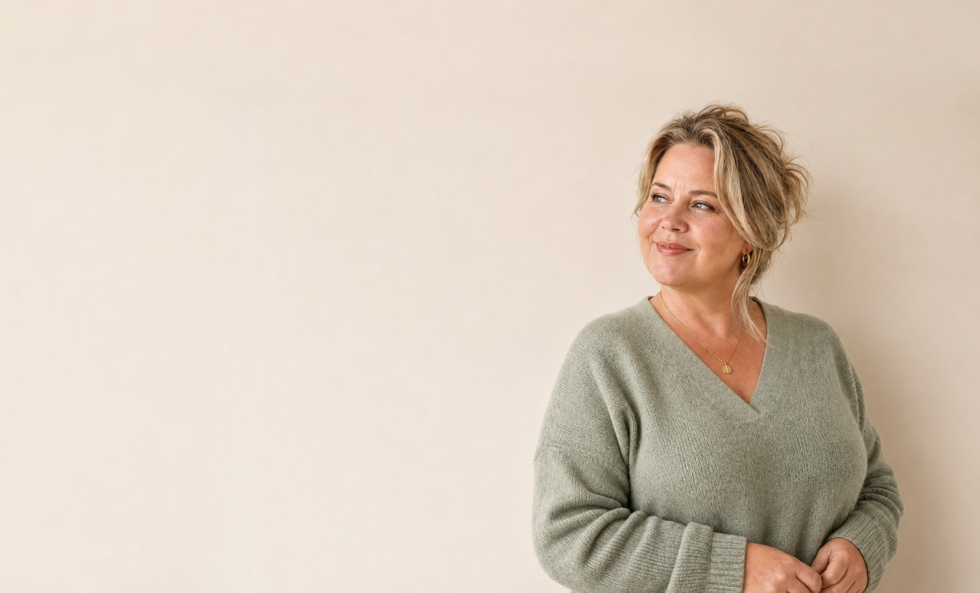 Vrouw van rond de vijftig in een groene trui glimlacht en kijkt opzij tegen een rustige beige achtergrond