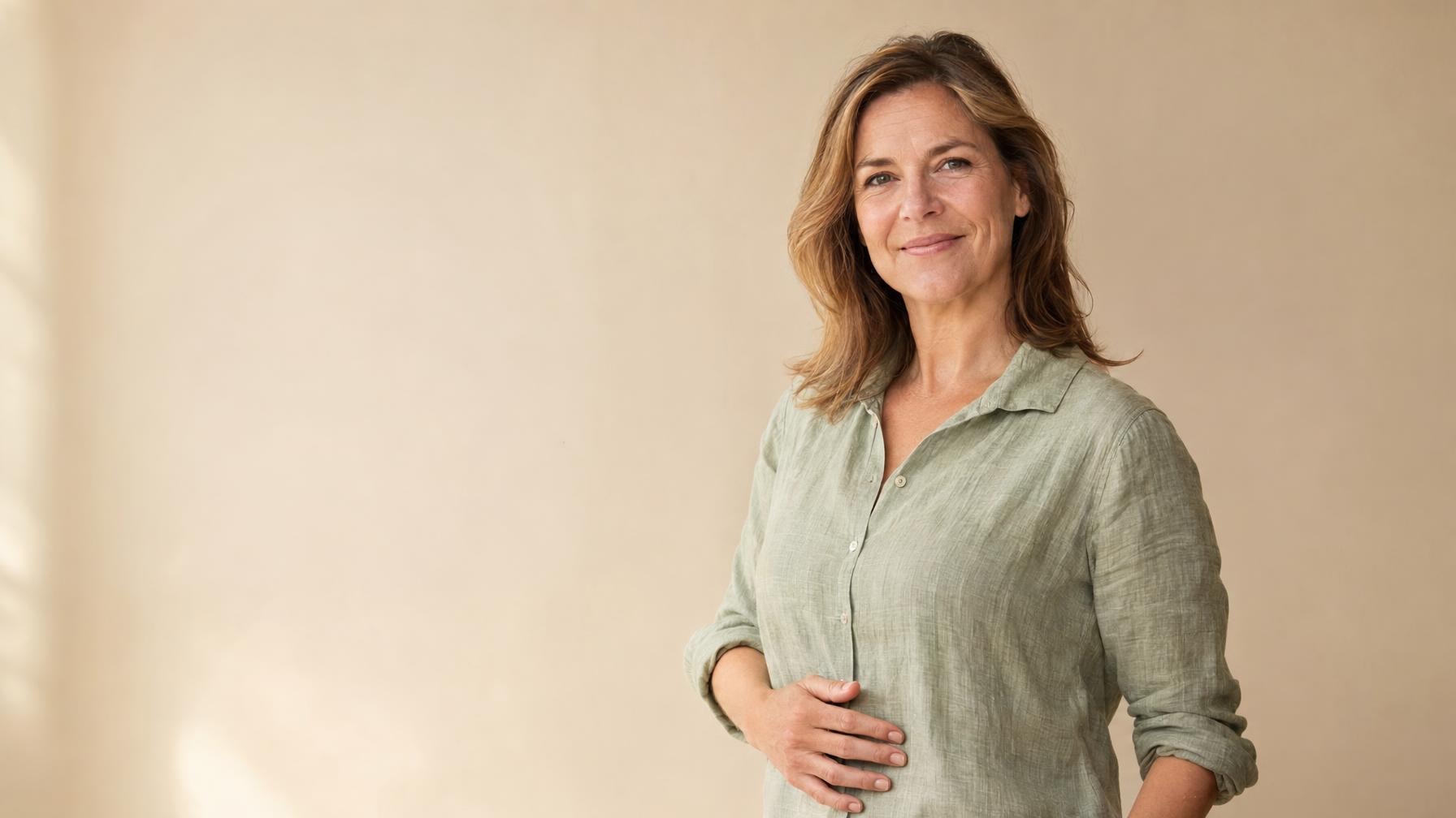 Vrouw van rond de vijftig in een linnen blouse met een hand op haar buik tegen een rustige beige achtergrond